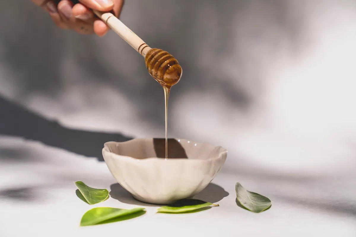 Golden honey dripping into a ceramic bowl beside green leaves